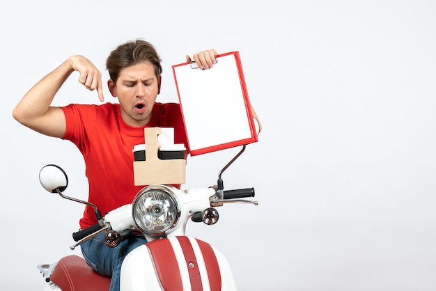 Jonge verwarde koeriersmens in rood uniform zittend op scooter met bestellingen en documenten op gele muur