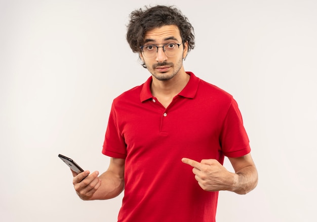 Jonge verwarde man in rood shirt met optische bril houdt en wijst op telefoon geïsoleerd op een witte muur