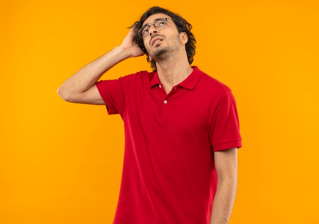Jonge verwarde man in rood shirt met optische bril houdt hoofd vast en kijkt omhoog geïsoleerd op oranje muur