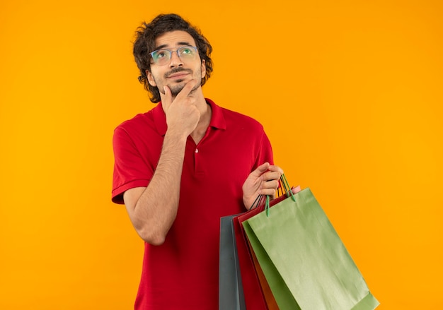 Jonge verwarde man in rood shirt met optische bril houdt veelkleurige papieren zakken en legt de hand op de kin geïsoleerd op een oranje muur