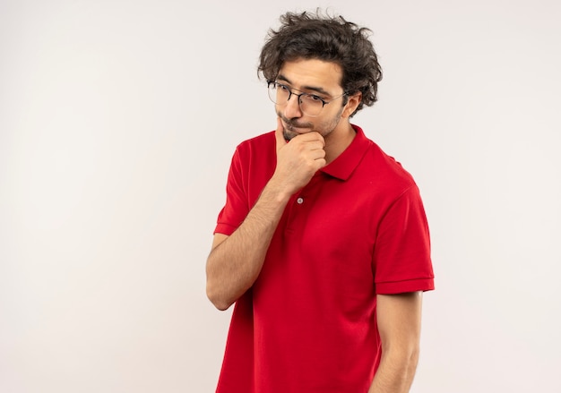 Jonge verwarde man in rood shirt met optische bril legt hand op kin en neerkijkt geïsoleerd op een witte muur