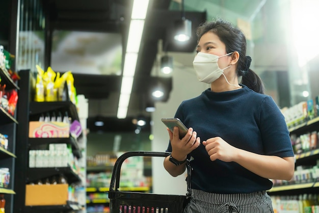 Jonge volwassen aziatische vrouwelijke vrouw die een beschermend gezichtsmasker draagt, die vers product of consumentenproduct kiest in het winkelcentrum van de kruidenierswinkel, met zorg en een vrolijke nieuwe normale winkellevensstijl