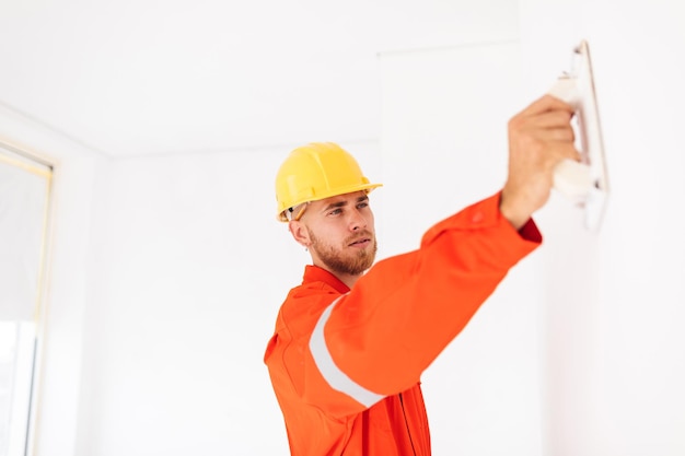 Gratis foto jonge voorman in oranje werkkleding en gele veiligheidshelm die zorgvuldig plamuurmes op het werk gebruikt