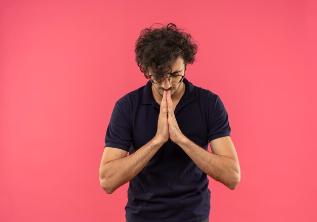 Jonge vreedzame man in zwart shirt met optische bril houdt handen bij elkaar en doet alsof ze mediteren geïsoleerd op roze muur