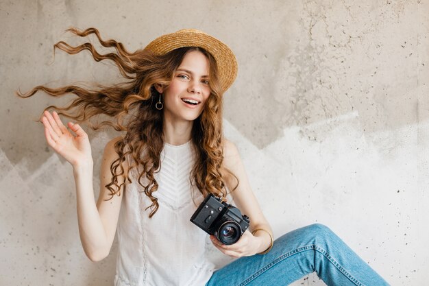 Jonge vrij glimlachende modieuze vrouw die blauwe denimjeans en wit overhemd draagt tegen muur in strohoed