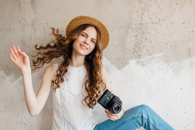 Jonge vrij glimlachende modieuze vrouw die blauwe denimjeans en wit overhemd draagt tegen muur in strohoed