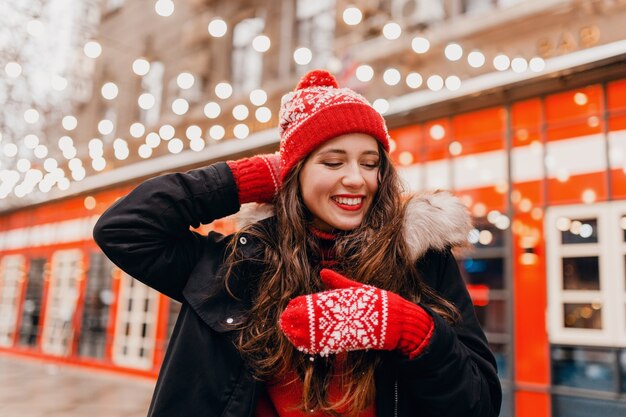 Jonge vrij lachende gelukkige vrouw in rode wanten en gebreide muts dragen winterjas wandelen in de stad straat, warme kleren