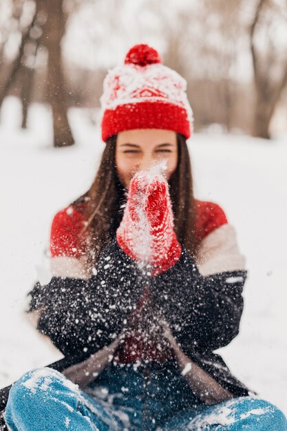 Jonge vrij lachende gelukkige vrouw in rode wanten en gebreide muts dragen winterjas, wandelen in het park, spelen met sneeuw in warme kleren