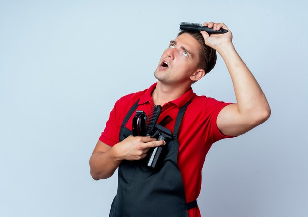 Jonge vrolijke blonde mannelijke kapper in uniform houdt kapper tools kammen van haar geïsoleerd op een witte ruimte met kopie ruimte