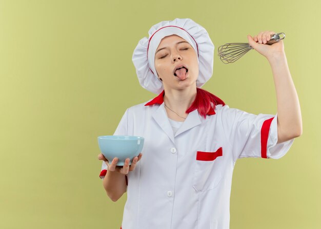 Jonge vrolijke blonde vrouwelijke chef-kok in chef-kok uniform stucks tong uit met gesloten ogen houdt kom en zwaait geïsoleerd op groene muur