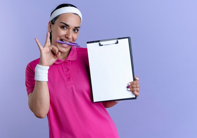 Jonge vrolijke Kaukasische sportieve vrouw met hoofdband en polsbandjes houdt pen met tanden gebaren ok handteken klembord houden op paars met kopie ruimte