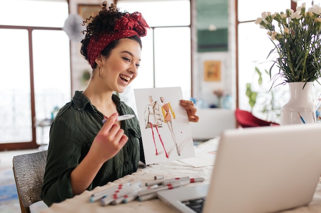 Gratis foto jonge vrolijke vrouw met donker krullend haar zittend aan tafel gelukkig mode schets in laptop tonen terwijl tijd doorbrengen in moderne gezellige werkplaats met grote ramen