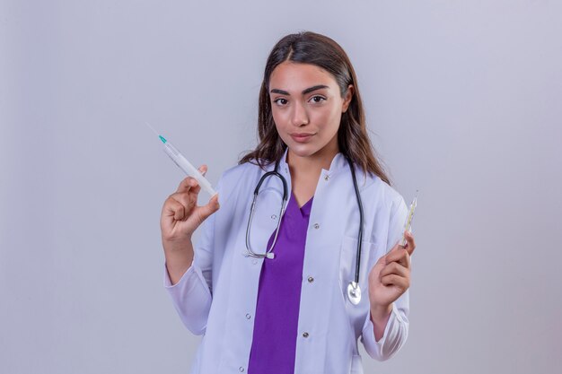 Jonge vrouw arts die in witte laag met phonendoscope de spuit van de cameraholding en thermometer met glimlach op gezicht over geïsoleerde witte achtergrond bekijken