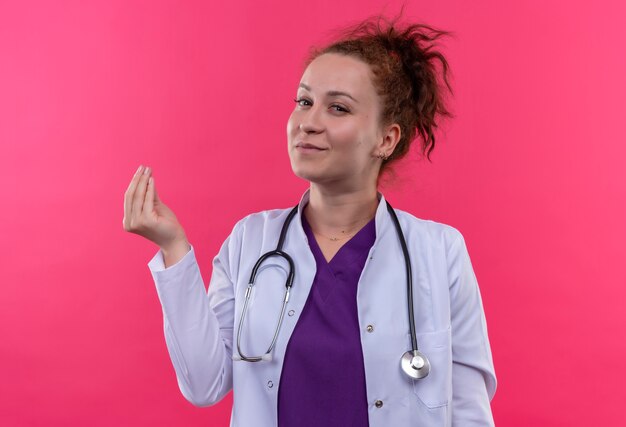 Jonge vrouw arts die witte jas draagt met een stethoscoop die vingers wrijft die geldgebaar maken met hand die zelfverzekerd om geld vragen die zich over roze muur bevinden