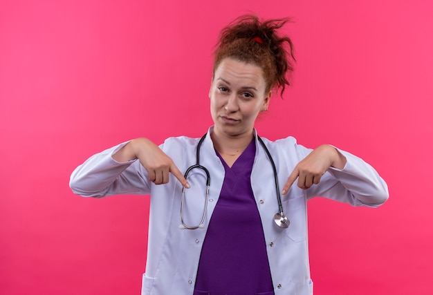 Jonge vrouw arts die witte jas met stethoscoop draagt die zelfverzekerd kijkt wijzend met indexfigers naar zichzelf staande over roze muur