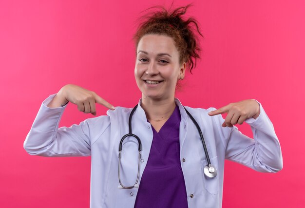 Jonge vrouw arts die witte laag met stethoscoop draagt die met vingers aan zichzelf richt die zich over roze muur bevinden