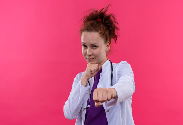 Jonge vrouw arts die witte laag met stethoscoop draagt die met vuist richt die zich zelfverzekerd over roze muur bevinden kijkt