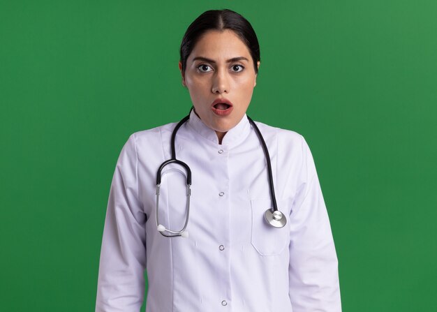 Jonge vrouw arts in medische jas met een stethoscoop om haar nek kijken naar voorkant verward en bezorgd staande over groene muur
