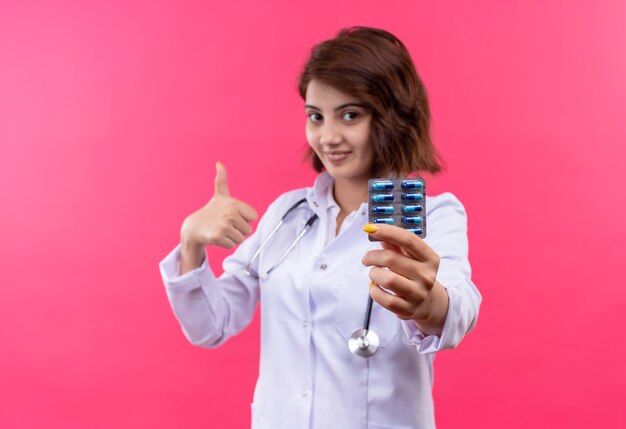 Jonge vrouw arts in witte jas met de blister van de stethoscoopholding met pillen die duimen opdagen glimlachen