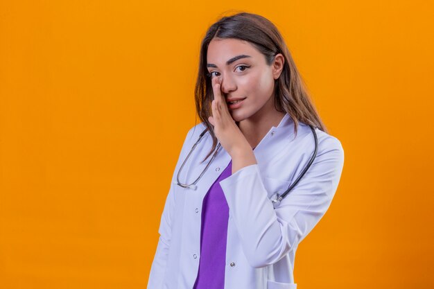Jonge vrouw arts in witte jas met phonendoscope die geheime informatie vertelt die haar hand houdt dichtbij mond over geïsoleerde oranje achtergrond