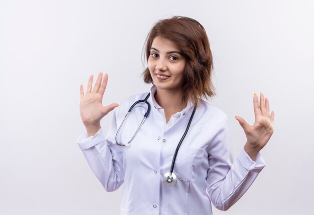 Jonge vrouw arts in witte jas met stethoscoop glimlachend verhogen handen in overgave staande over witte muur