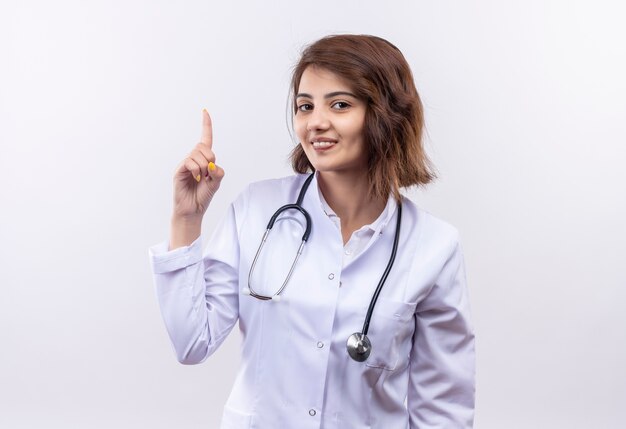 Jonge vrouw arts in witte jas met stethoscoop glimlachend vertrouwen tonen wijsvinger met nieuw geweldig idee staande over witte muur