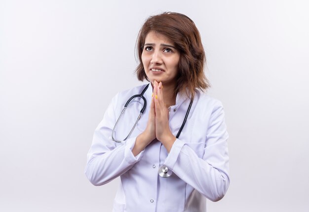 Jonge vrouw arts in witte jas met stethoscoop hand in hand in gebed gebaar, bedelen met hoop expressie, bezorgd