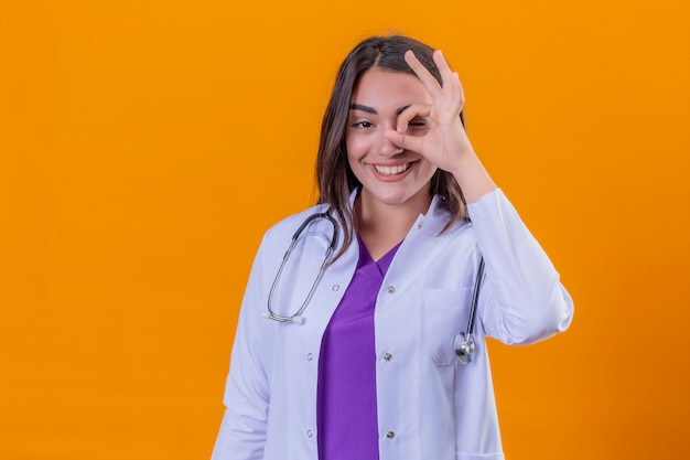 Jonge vrouw arts in witte laag met phonendoscope die ok teken met hand op oog doet die door vingers met gelukkig gezicht over geïsoleerde oranje achtergrond kijkt