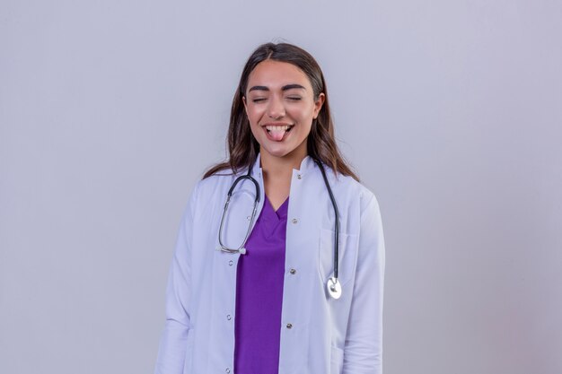 Jonge vrouw arts in witte laag met phonendoscope grappige en vriendschappelijke uitstekende tong die zich op wit geïsoleerde achtergrond bevinden