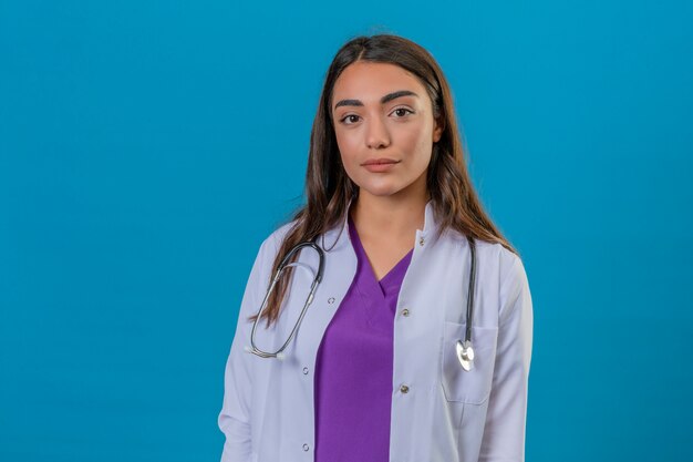 Jonge vrouw arts in witte laag met phonendoscope met ernstig gezicht dat zich op blauw geïsoleerde achtergrond bevindt
