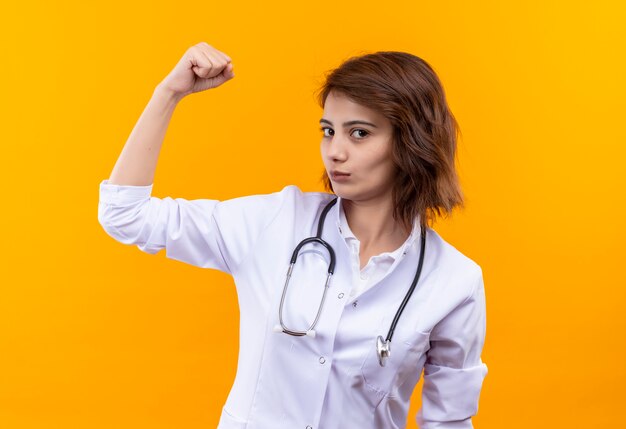 Jonge vrouw arts in witte laag met stethoscoop die vuist opheft die biceps toont die zich zelfverzekerd over oranje muur bevinden kijken