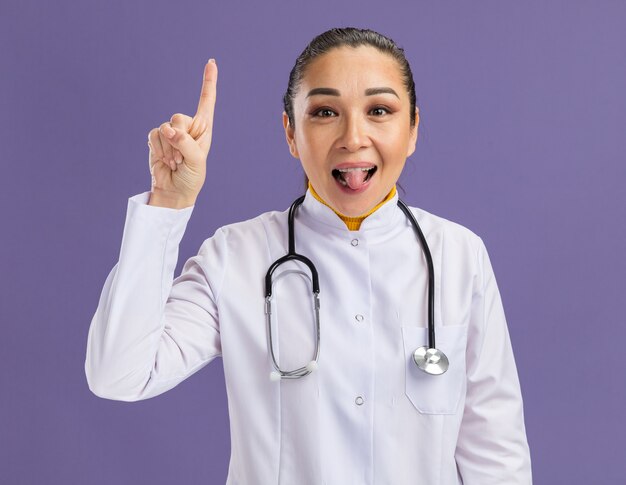 Jonge vrouw arts in witte medicijnjas met stethoscoop om nek blij en verrast met wijsvinger die tong uitsteekt die over paarse muur staat