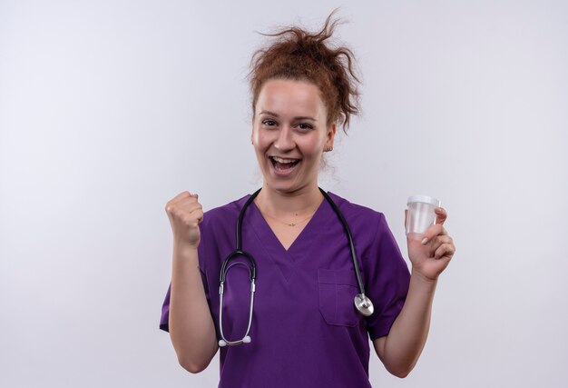 Jonge vrouw arts medische uniform dragen met een stethoscoop houden testpot blij en verlaten balde vuist staande over witte muur