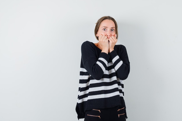 Jonge vrouw balde vuisten en hand in hand op de mond in gestreepte gebreide kleding en zwarte broek en bang op zoek