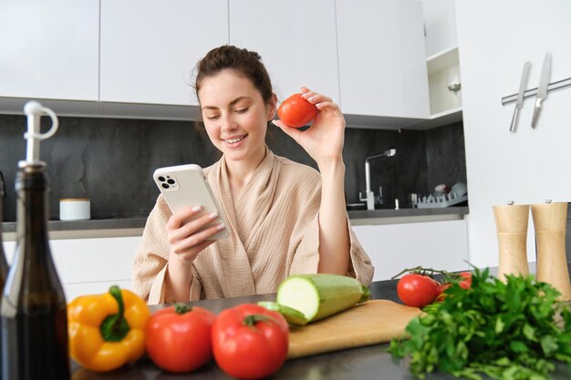 Jonge vrouw bestelt boodschappen via de mobiele app. Meisje in badjas zit in de keuken met groenten