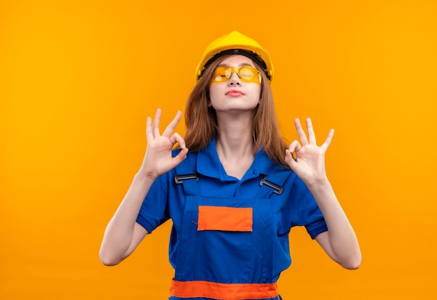 Jonge vrouw bouwer werknemer in bouw uniform en veiligheidshelm staande met gesloten ogen ontspannen meditatie gebaar met vingers over oranje muur maken