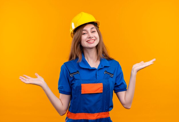 Jonge vrouw bouwer werknemer in de bouw uniform en veiligheidshelm op zoek spreidende armen naar de zijkanten glimlachend staande