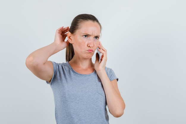 Jonge vrouw denken terwijl praten over mobiel in grijs t-shirt en somber op zoek. vooraanzicht.