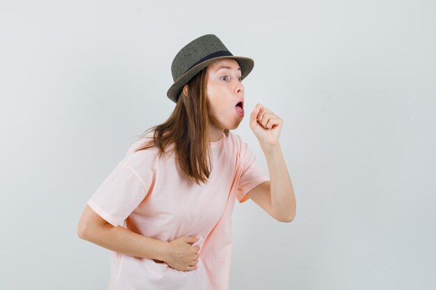 Jonge vrouw die aan hoest in roze t-shirt, hoed lijdt en ziek, vooraanzicht kijkt.