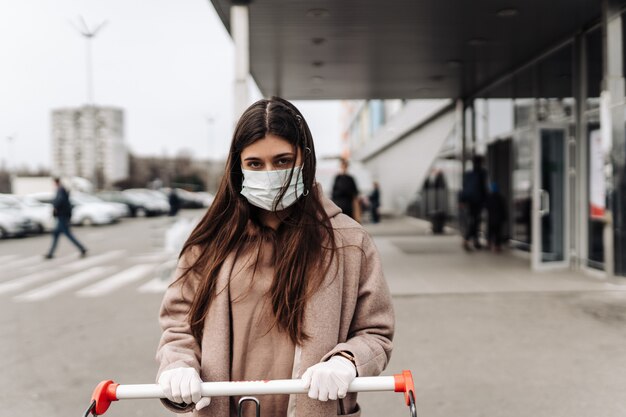Jonge vrouw die beschermingsgezichtsmasker draagt tegen coronavirus 2019-nCoV die een boodschappenwagentje duwt.