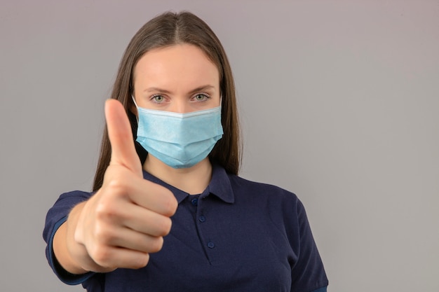 Jonge vrouw die blauw poloshirt in beschermend medisch masker draagt dat duim op positieve uitdrukking toont die zich op lichtgrijze achtergrond bevindt