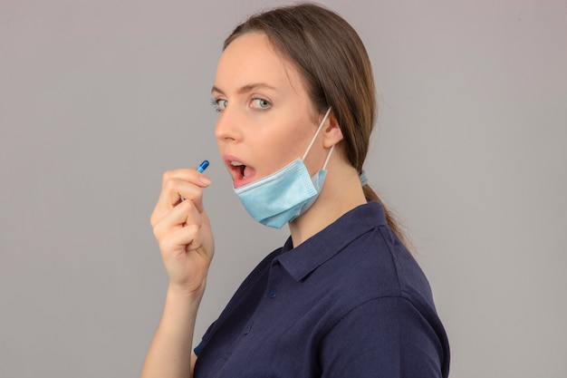 Jonge vrouw die blauw poloshirt in beschermend medisch masker met open mond draagt die een pil op lichtgrijze achtergrond neemt