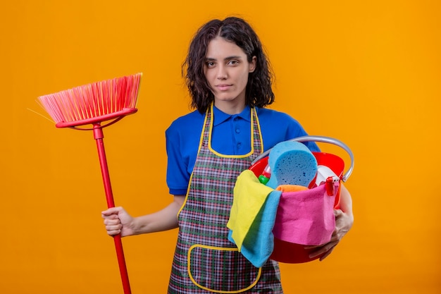 Jonge vrouw die de emmer van de schortholding met het schoonmaken van hulpmiddelen en dweil draagt ?? die camera met ernstige zekere uitdrukking bekijkt die zich over oranje achtergrond bevindt