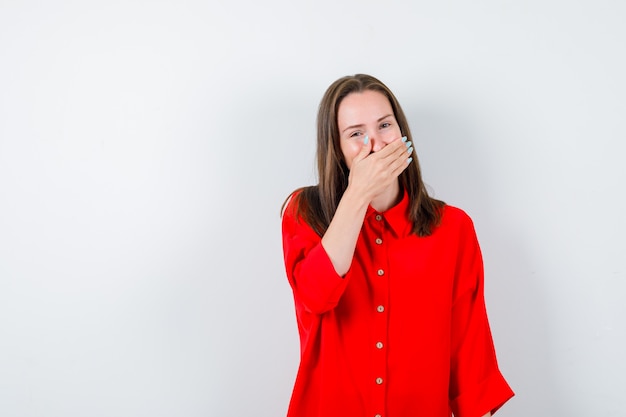 Jonge vrouw die de hand op de mond houdt in een rode blouse en er vrolijk uitziet, vooraanzicht.