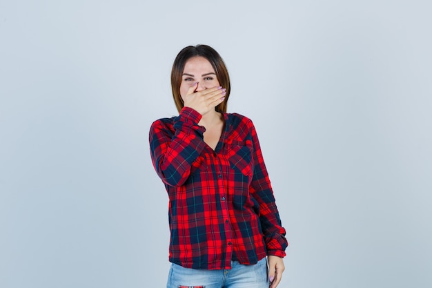 Jonge vrouw die de mond bedekt met de hand terwijl ze lacht in een geruit overhemd, een spijkerbroek en er vrolijk uitziet, vooraanzicht.