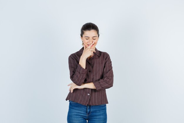 Jonge vrouw die de mond bedekt met de hand terwijl ze lacht in een gestreept shirt, spijkerbroek en er vrolijk uitziet, vooraanzicht.