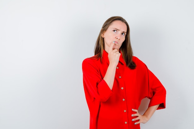 Jonge vrouw die de vinger op de rand van de lippen houdt, omhoog kijkt in een rode blouse en peinzend kijkt, vooraanzicht.