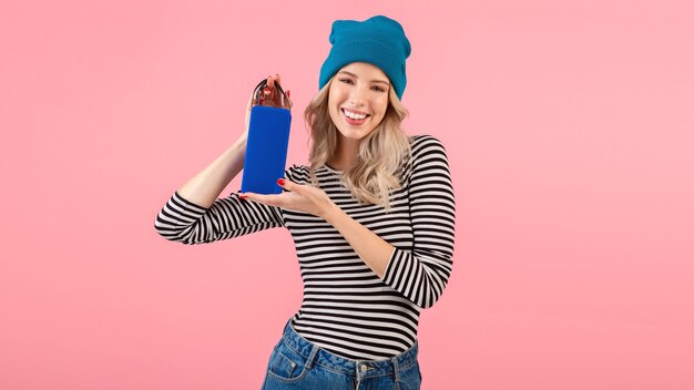 Jonge vrouw die draadloze spreker houdt die aan muziek luistert die een gestreept overhemd en een blauwe hoed draagt die die zich op roze stelt glimlachend