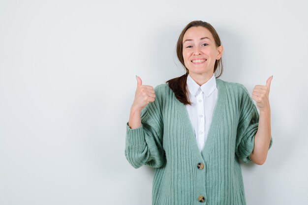 Jonge vrouw die dubbele duimen in blouse, vest toont en er vrolijk uitziet, vooraanzicht.