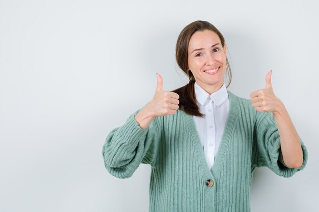 Jonge vrouw die dubbele duimen in blouse, vest toont en er vrolijk uitziet, vooraanzicht.